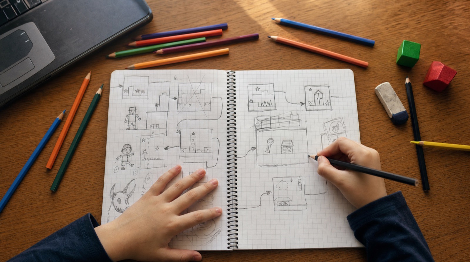Vista cenital de un cuaderno con el plan de un videojuego dibujado a lápiz: pantallas, flechas e ideas tachadas, con las manos de un niño trabajando, lápices de colores y un portátil al borde de la mesa.